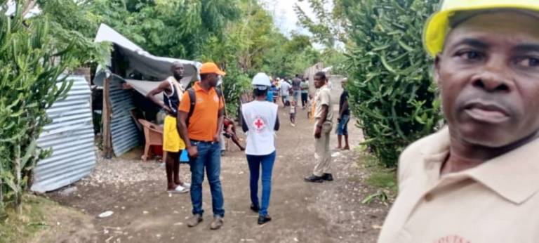 man in Red Cross vest and others in hard hats walk down a dirt road