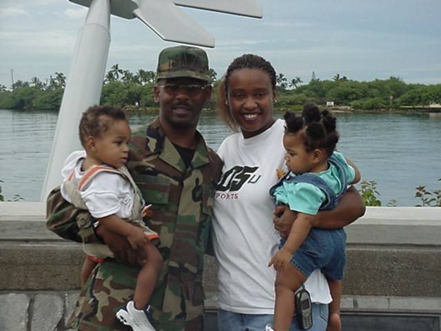 An old family photo of a man in a full Army uniform and hat holds one child while his wife holds the other. They smile with a river in the background.