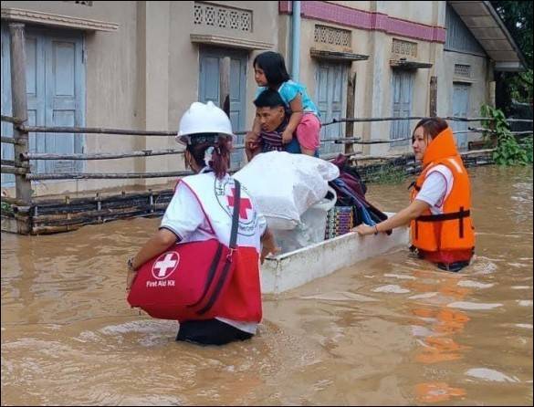 Un hombre y una mujer con chalecos de la Cruz Roja empujando una pequeña embarcación a través de las aguas de inundación llevando a un hombre, un niño pequeño y sus pertenencias.