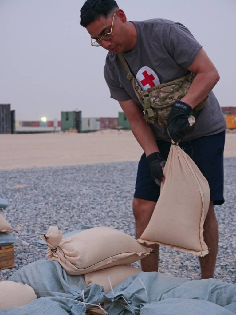 Soldier with Red Cross shirt heaves a sandbag besides several others in a desert.