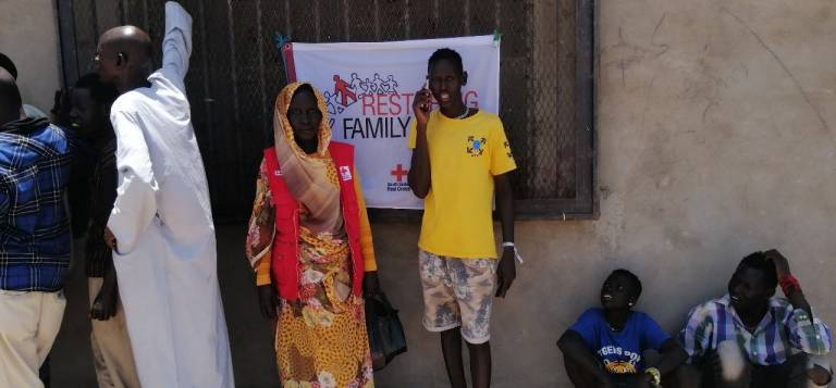 Man stands, talking on a phone while woman in Red Cross vest looks on.