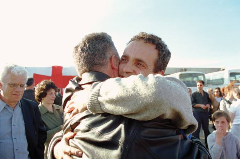 Two men hug with other people in the background watching.