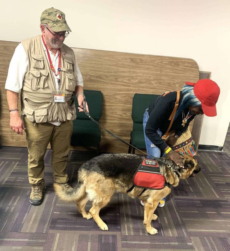 Man in Red Cross vest with German Shepherd service dog and woman petting the dog.