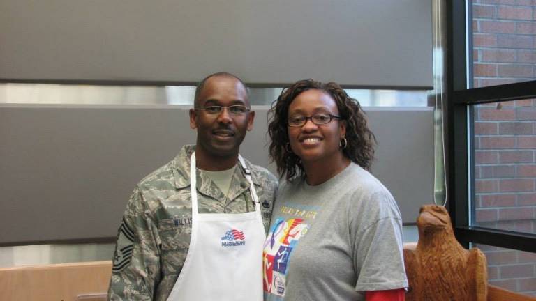 A man in his Air Force uniform, a shaved head and an apron smiles besides his wife. 