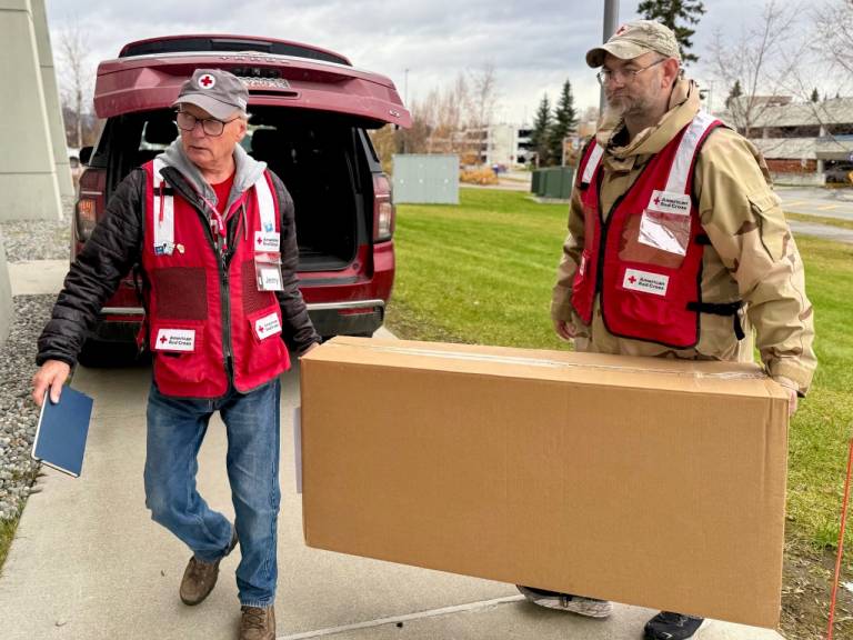 Two men in Red Cross vests carry supplies for a shelter