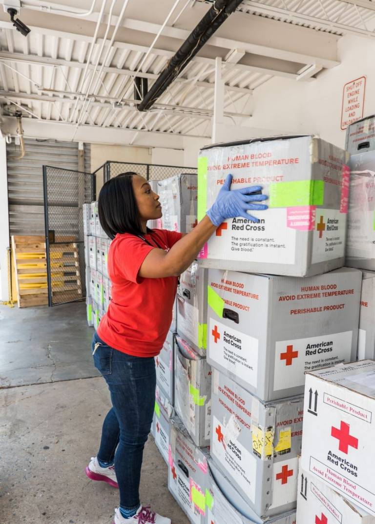 woman in Red Cross shirt standing outside van packed with blood products ready to be delivered to hospitals.