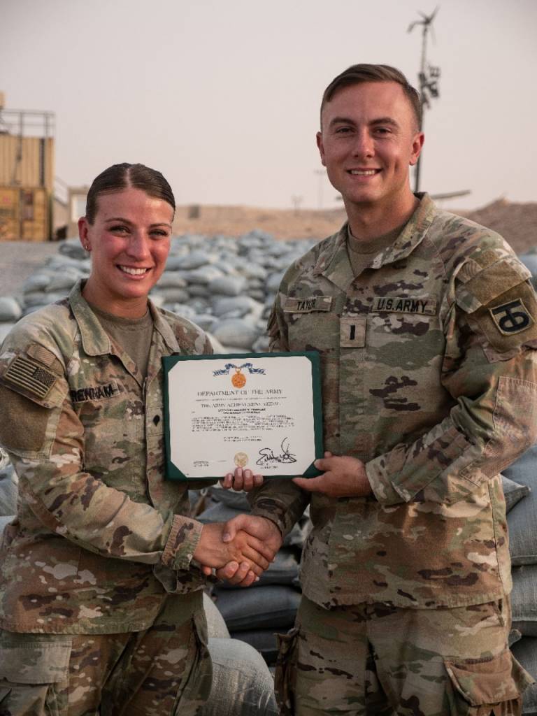 Heather holds a Military Outstanding Volunteer Service Medal award certificate, smiling beside her commanding officer and in front of all the sand bags. 