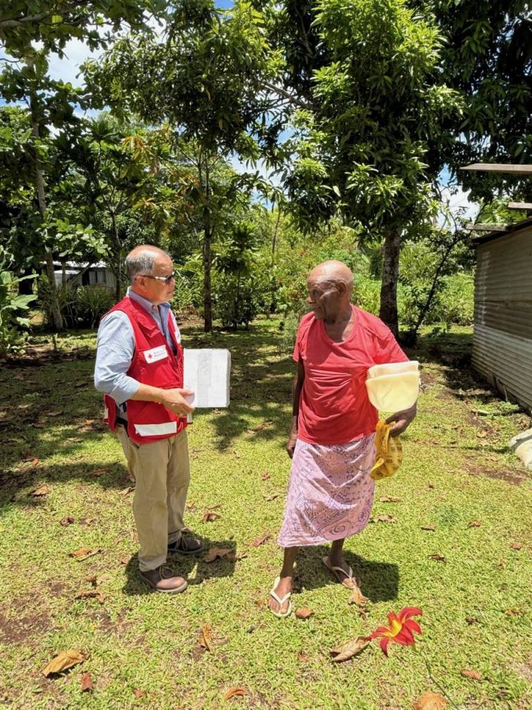 Two men stand talking in the sunshine with packages in their arms. 