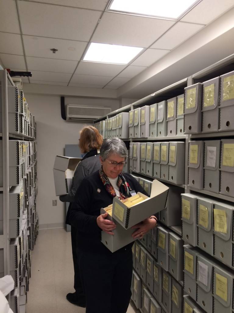 Two older women look through binders and folders in the vault of the Red Cross archives in Washington, D.C. The space is small, brightly lit and windowless. 