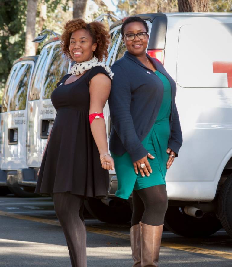 Two women standing back to back and smiling in front of an American Red Cross vehicle. The woman on the left has a red donation bandage on her arm.