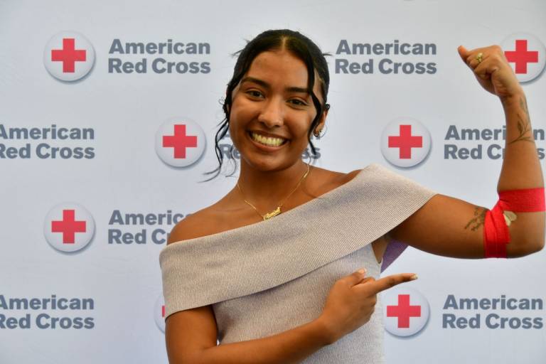 A young woman smiling and pointing to a red donation bandage on her arm while standing in front of an American Red Cross step-and-repeat banner.