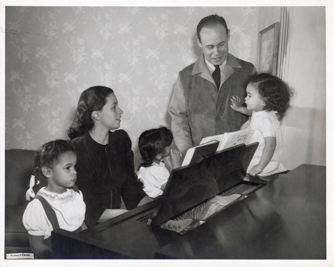 Dr. Charles R. Drew stands smiling at his wife Lenore and three young daughters gathered around a piano in their family home.