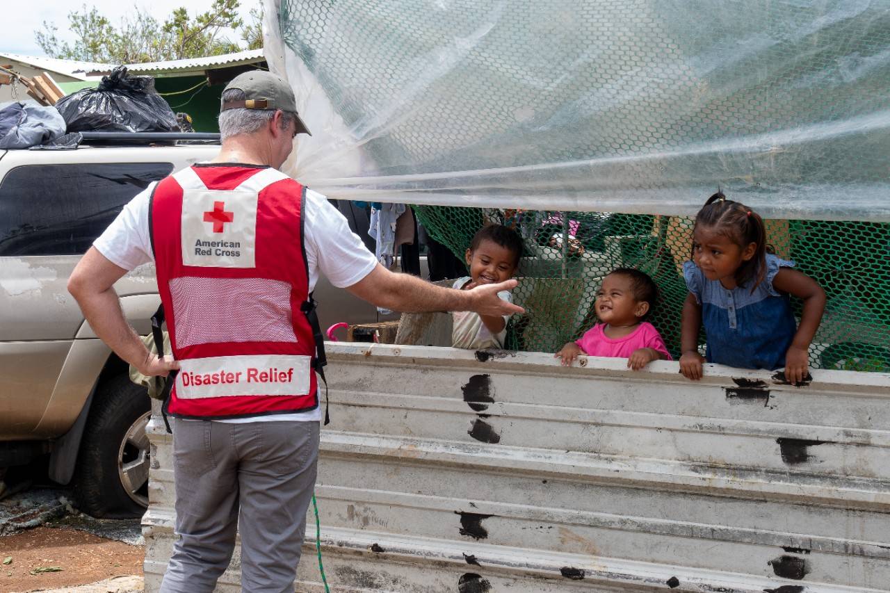 Man in Red Cross vest amuses three small children. 