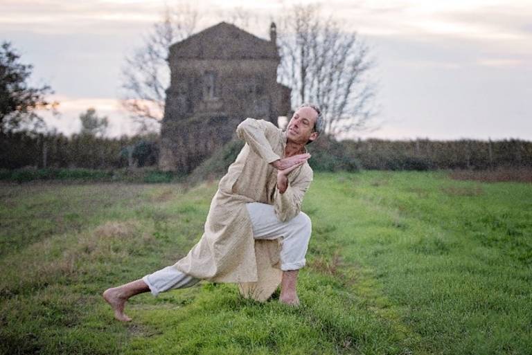 Doug lunges forward and twists at the waist. His hands are in prayer pose. He wears loose-fitting clothing, barefoot in the grass. Behind him is a field and what looks like an out-of-focus farmhouse.