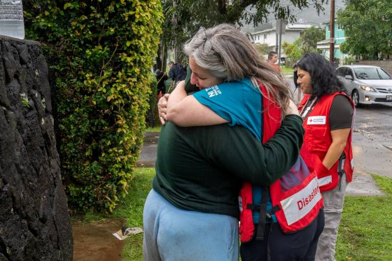 Three women, two in Red Cross vests, one of which is hugging a woman trying to clean up after the flooding. 