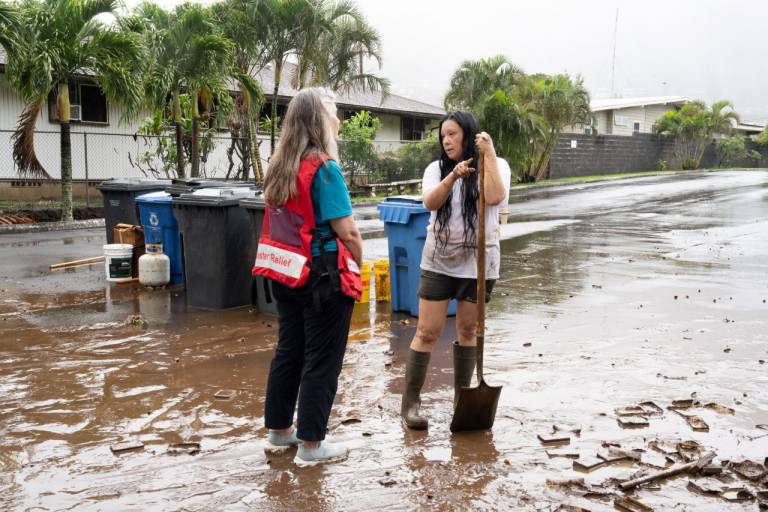 Woman in Red Cross vest speaks with woman leaning on a shovel