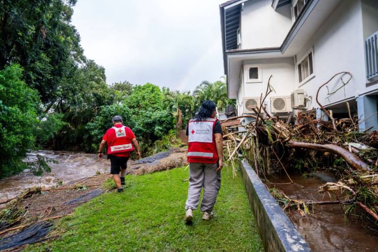 Two people in Red Cross vests walk through debris left behind by flooding