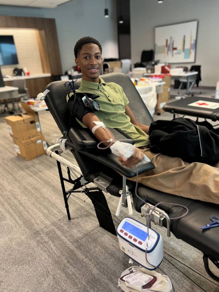Grant Buckley, a young Black man wearing a green polo shirt, smiles at the camera while donating blood at a Red Cross blood drive.