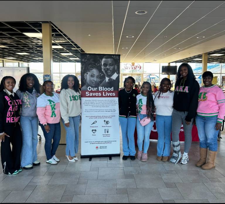 A group of nine young Black women wearing Alpha Kappa Alpha Sorority, Incorporated apparel in pink and green stand together at a blood drive, flanking an American Red Cross banner that reads "Our Blood Saves Lives."