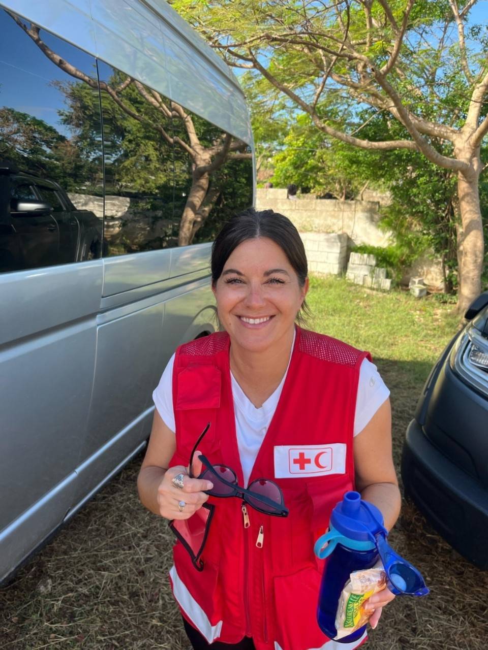 A woman in a red vest stands smiling in front of a van. 
