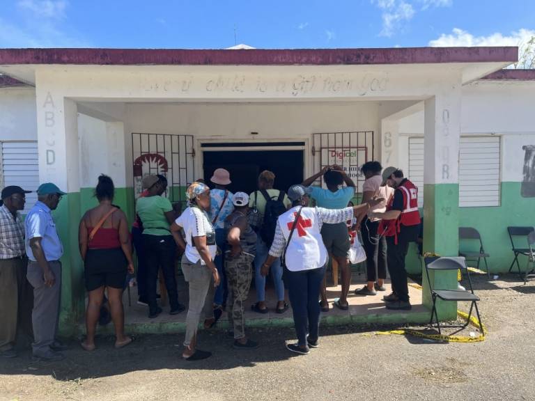 A group of people gather in the sunshine outside the doors of a community center in Jamaica. 