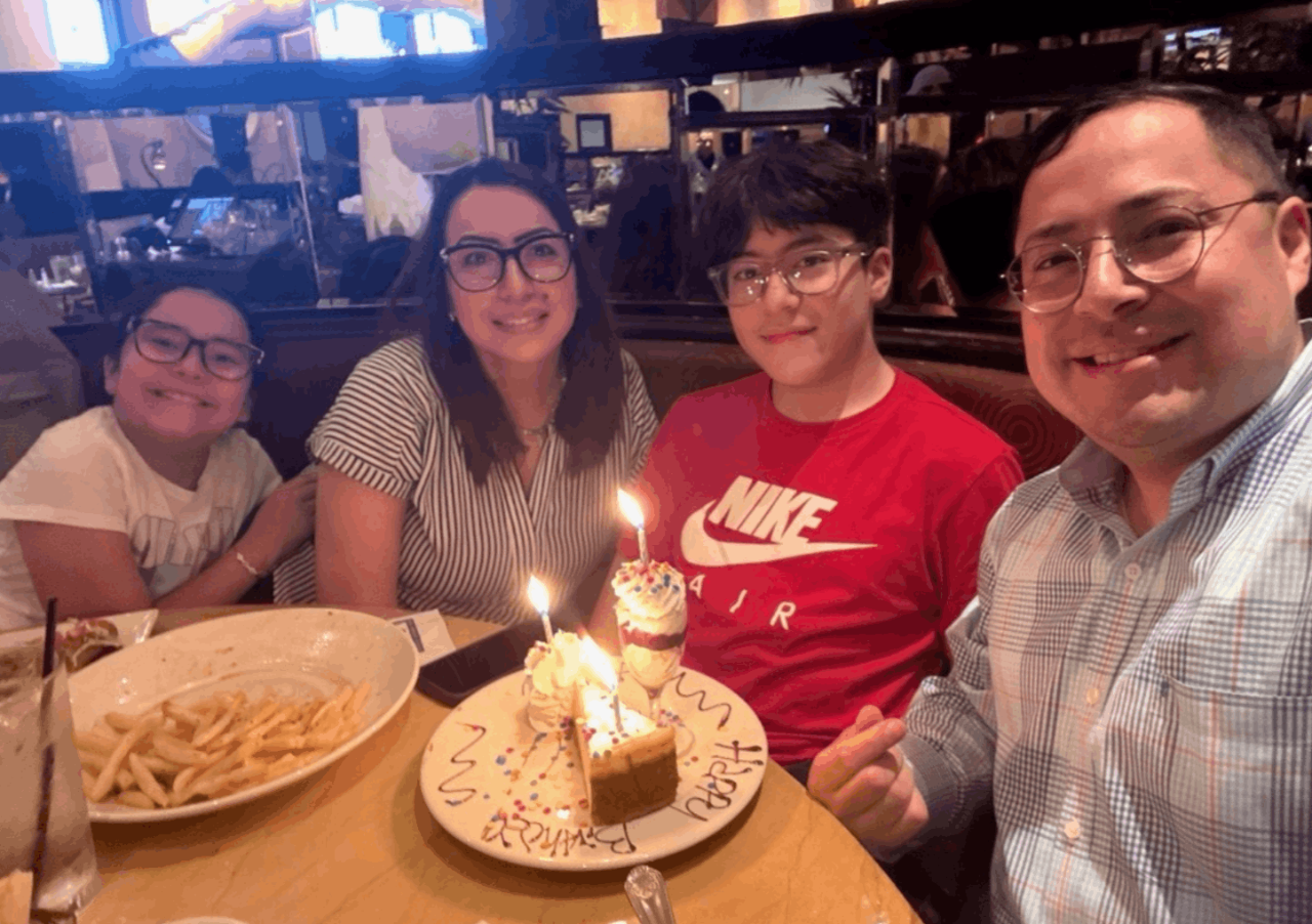 Familia de cuatro personas sentadas en torno a una mesa en donde hay un plato con papas fritas y un pastel con una vela en un plato en donde se lee: Happy Birthday. Sentados de izquierda a derecha: niña, mamá, niño, papá