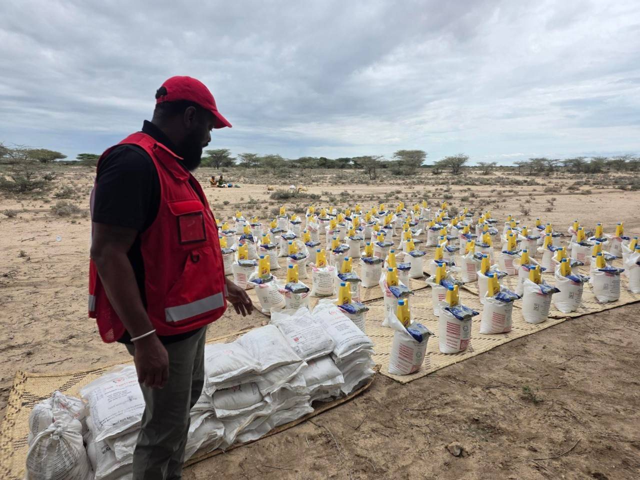 A man in a red vest and red baseball cap stands in front of a tarp with stacks of agricultural supplies ready for distribution in the local community. Trees and dry terrain can be seen in the background. 