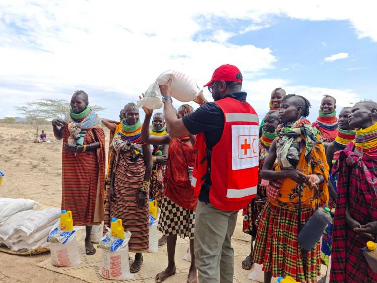 A man in a Red Cross vest, a red ball cap and tan pants hands a bag of dry goods to a community member. Other members of the community gather around, all wearing brightly colored cultural attire. Trees and dry terrain can be seen in the background. 