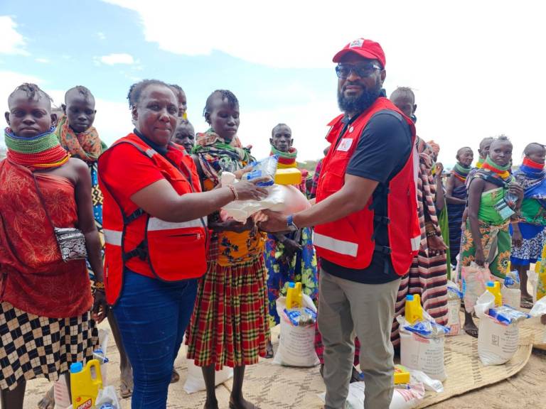 Two individuals in red vests hand dry goods and agricultural supplies to community members gathered nearby, all wearing brightly colored cultural attire. Stacks of supplies sit on the ground, arranged in rows.