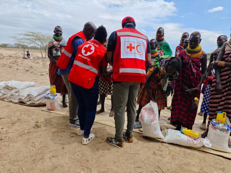 Three individuals in red vests hand dry goods and agricultural supplies to community members gathered nearby. Community members are all wearing brightly colored cultural attire. Stacks of supplies sit on the ground, arranged in rows, and trees and dry terrain can be seen in the background. 