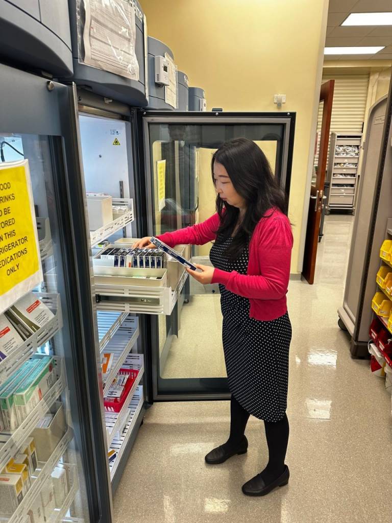 A woman in a red sweater and a black dress stands beside a glass cabinet, sorting medications in a drawer. 