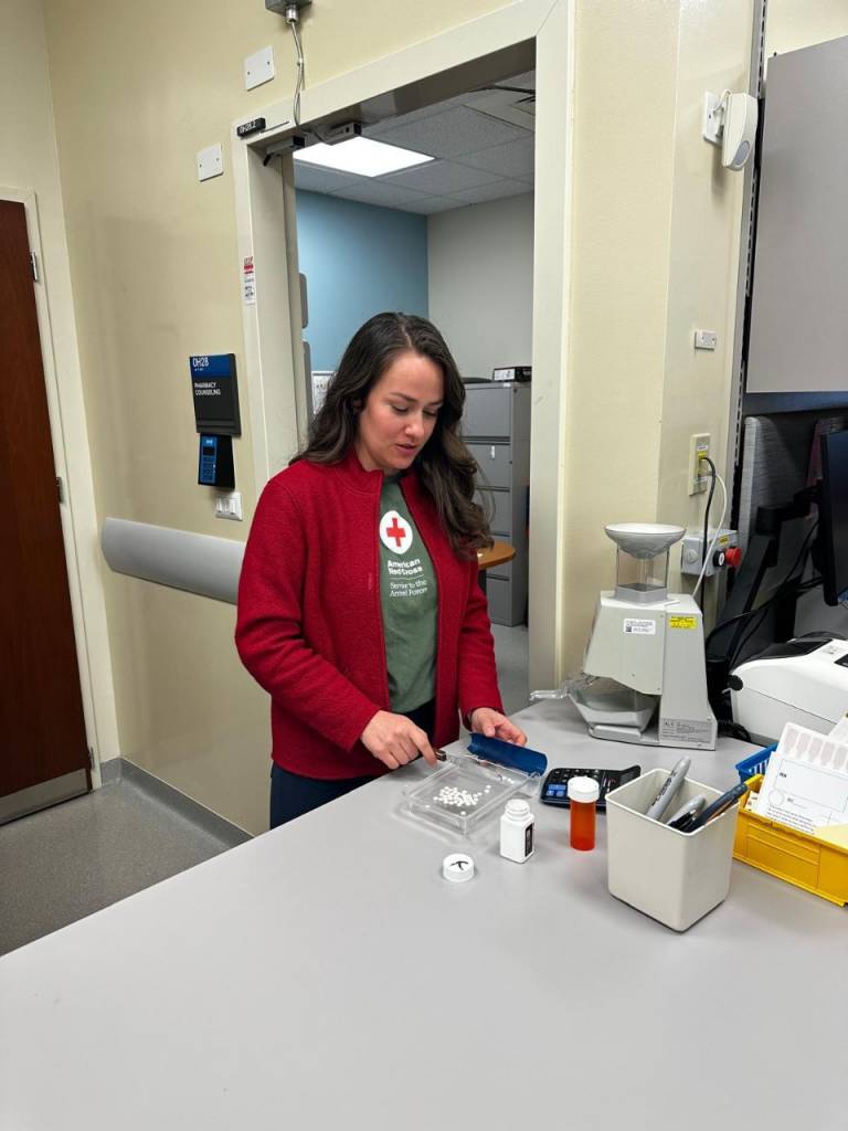 A woman in a red sweater and a green Red Cross t-shirt stands in front of a counter and sorts small white pills.