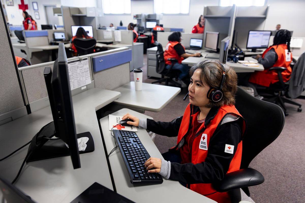 Woman in Red Cross vest sits at desk looking at a computer.