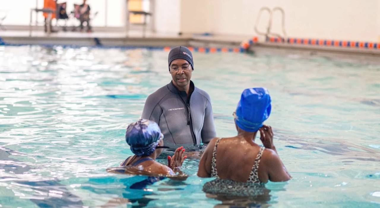 A person standing in an indoor swimming pool with a swim cap on talking to two other swimmers who are both wearing swim caps. 
