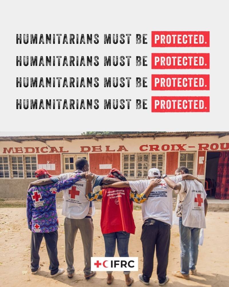 A group of five people in Red Cross clothing on stand in a row with their backs to the camera and their arms wrapped around each other’s shoulders. A rural medical building can be seen in the background.