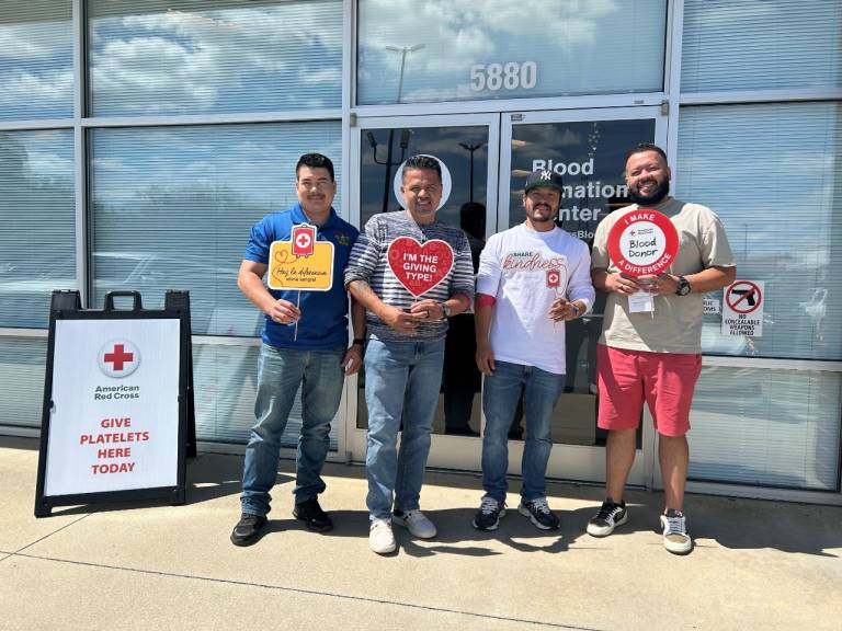 Four men stand outside a Red Cross blood donation facility holding blood donor signs.