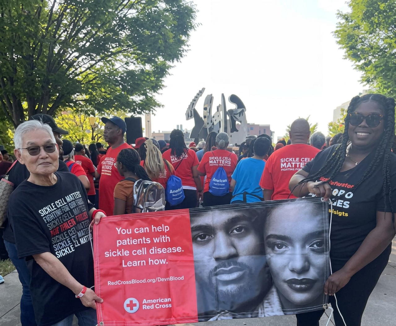 Two people hold a red American Red Cross banner reading "You can help patients with sickle cell disease. Learn how." at an outdoor walk.