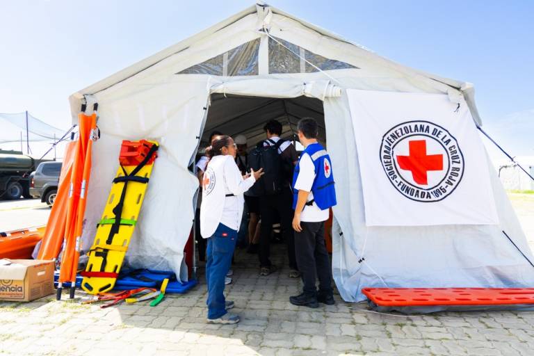 Multiple people stand in the entryway of a white makeshift medical tent with the Red Cross logo displayed prominently on the outside. A stretcher leans up against the wall of the tent. 