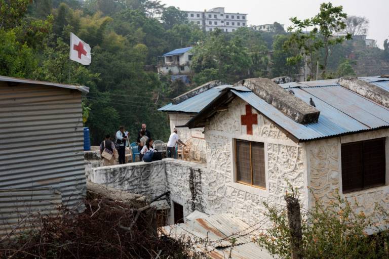A white building with a Red Cross symbol on the side of it can be seen among hills and green trees.