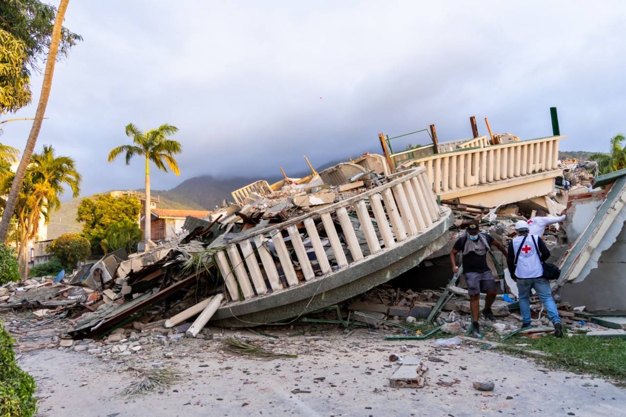 Three people, one in a black shirt, mask and baseball cap and two with Red Cross vests can be seen climbing amongst rubble from the earthquake. 