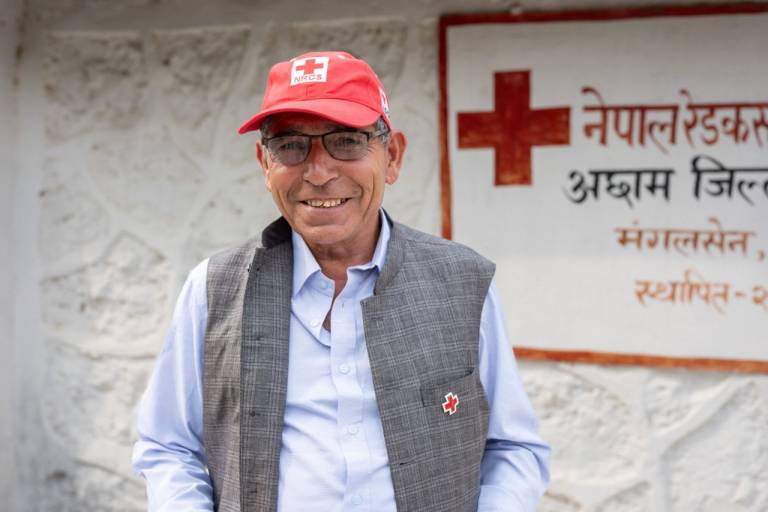 A man wearing a Red Cross ball cap is seated at a desk and gestures with his hands while having a conversation.
