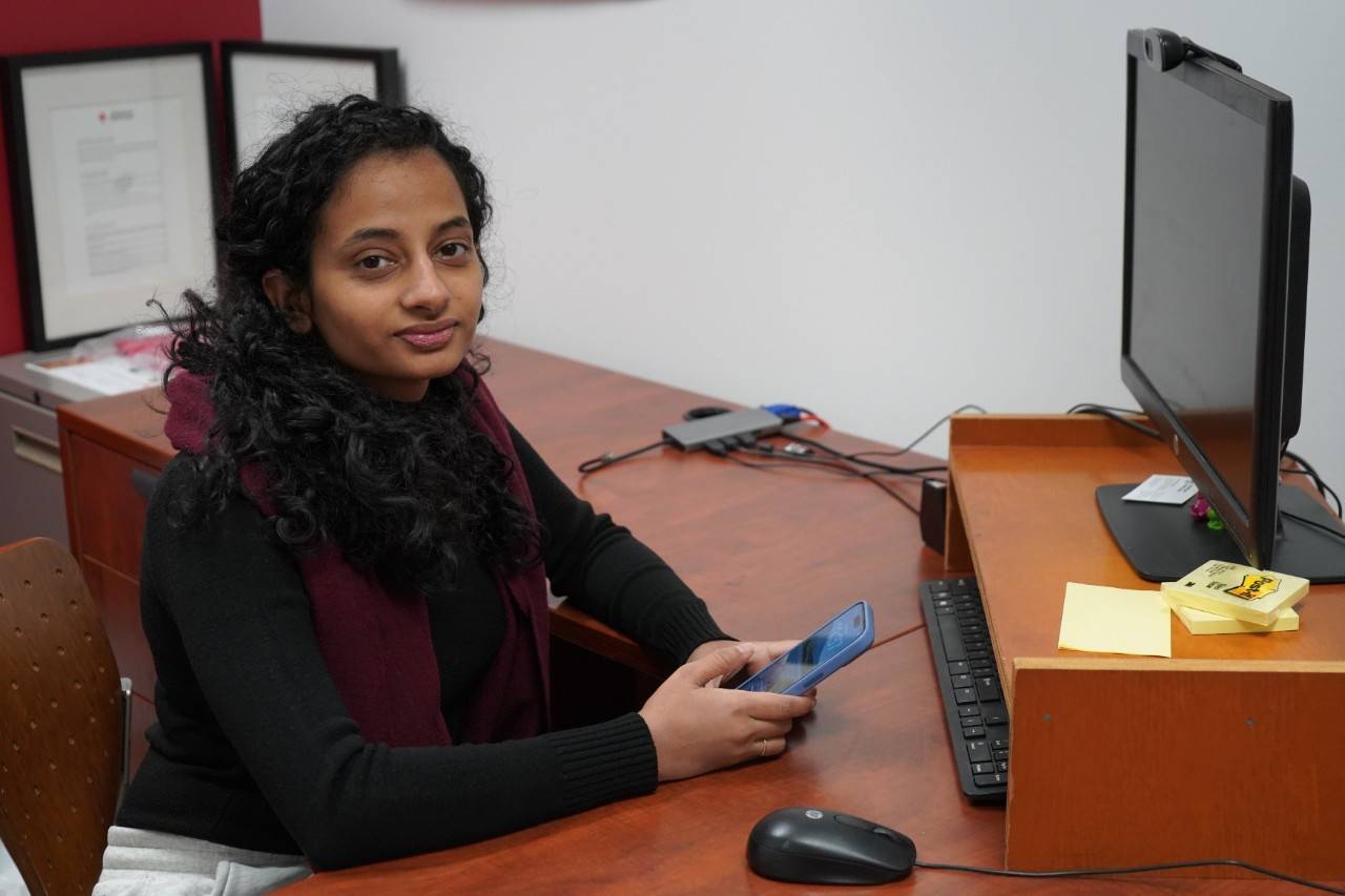Una mujer con una camisa negra y una bufanda color granate, sentada en un escritorio y sosteniendo un teléfono celular. 