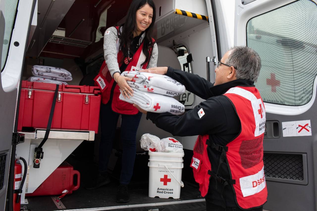 Woman in Red Cross vest hands blankets out of a vehicle to a man also in a Red Cross vest.