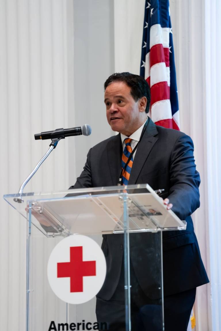 A man in a dark suit and striped tie speaks at a clear podium bearing the American Red Cross logo, with a U.S. flag behind him.