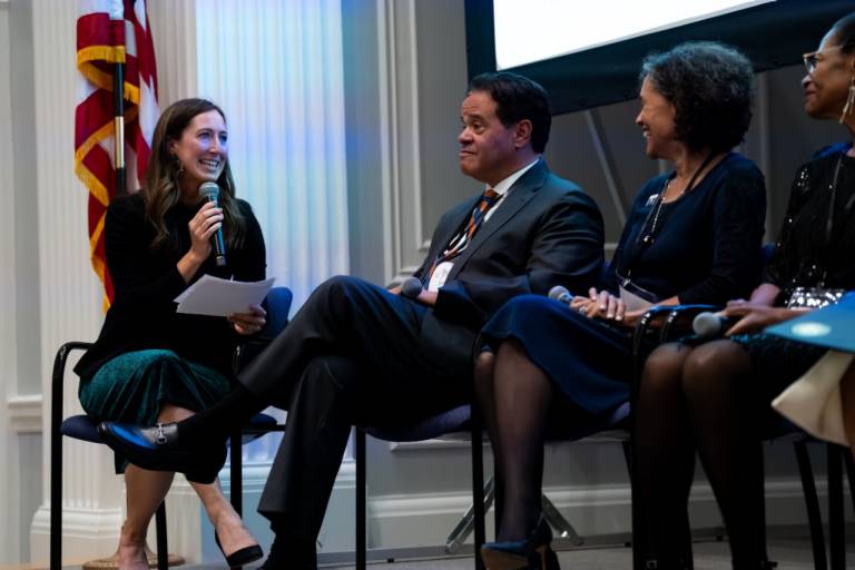 A woman holding a microphone and notes laughs while speaking to three seated panelists beside her, with a U.S. flag in the background.
