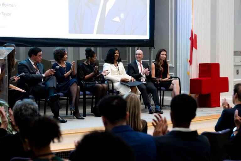 Six panelists seated on a stage applaud as an audience watches, with a large red cross sculpture and a projected image of Dr. Drew behind them.