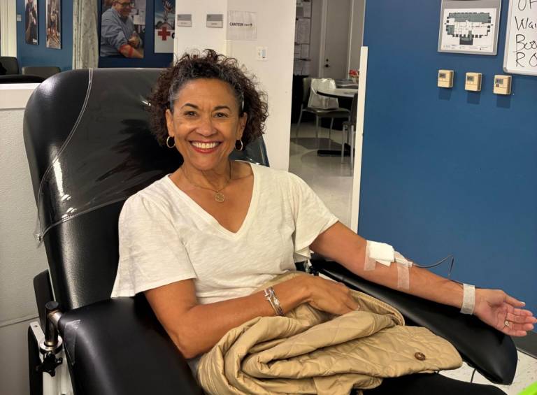 A smiling woman in a white shirt reclines in a black donor chair with a bandaged arm extended, a blanket in her lap and donor posters on the wall behind her.