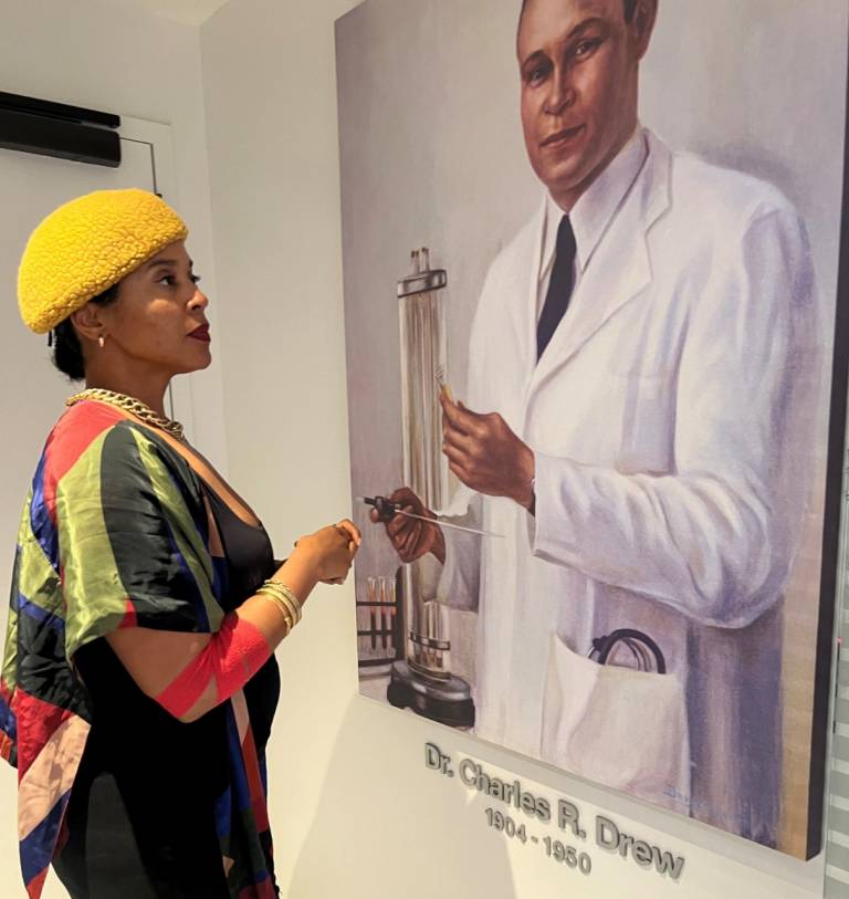 A woman in a yellow hat and colorful striped wrap gazes up at a painted portrait of Dr. Charles R. Drew in a white lab coat, labeled with his name and the years 1904 to 1950.