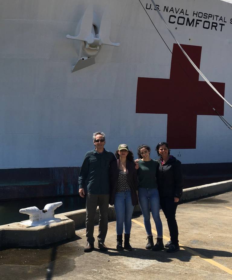 Doug, his wife and their two daughters take a photo beside the U.S. Naval Hospital Ship Comfort. The ship is too large to fit in the frame, but a few mooring lines and a large anchor off the side of the ship can be seen. 