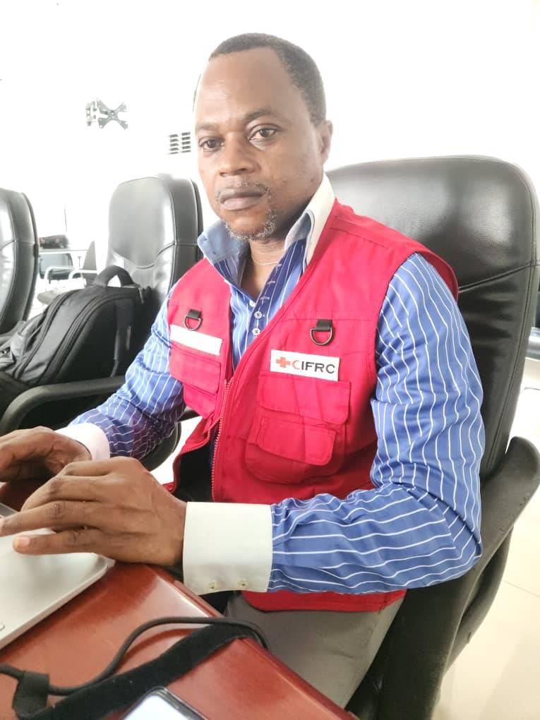A man in a red vest and a blue button-down shirt sits at a desk and types on a computer keyboard.
